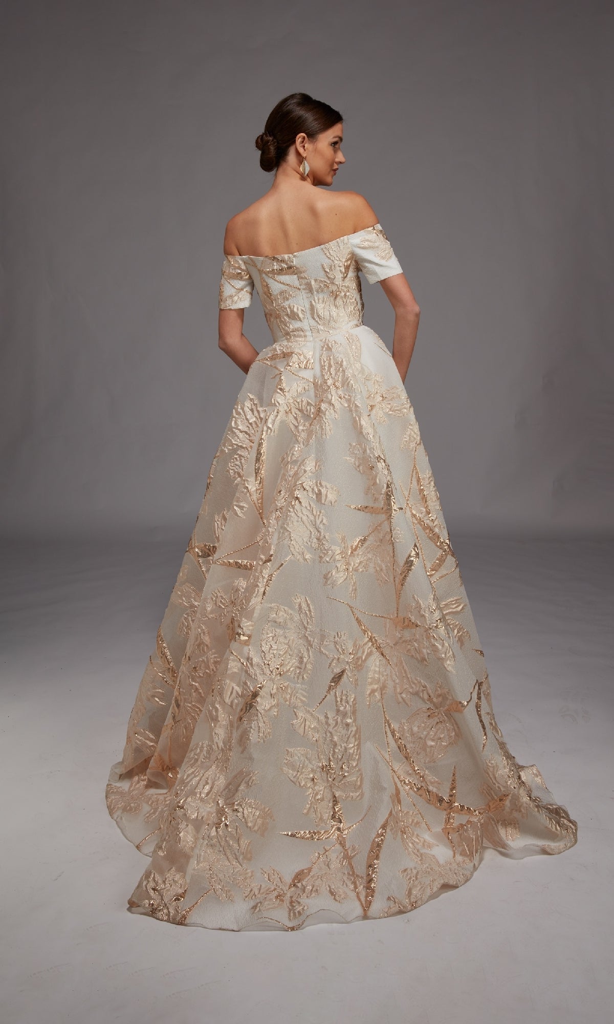 A woman faces away, wearing the Alyce 27804 long formal dress—an off-the-shoulder, floor-length gown featuring gold floral embroidery and metallic leaf motifs on a sheer overlay.